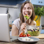 young woman eating a salad in front of laptop in kitchen