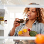 View Looking Out From Inside Of Refrigerator As Woman Opens Door And Packs Food Onto Shelves