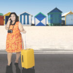 woman using a mobile phone while standing with a suitcase in front of beach cottages