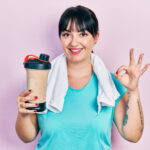 Young hispanic woman wearing sport clothes drinking a protein shake doing ok sign with fingers